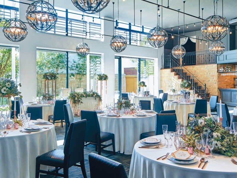 高い天井から吊り下げられたシャンデリアが印象的な、開放感あふれるパーティ会場です。大きな窓から自然光が差し込み、白いテーブルクロスと花のテーブル装飾が上品な雰囲気を演出しています。ウェディングや各種パーティに最適な、洗練された空間です。