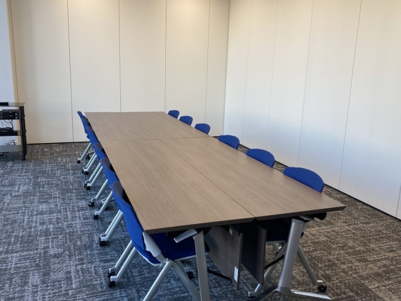 木目調テーブルとブルーチェアを整然と並べたスクール形式のレイアウト。最大12名まで着席可能で、シンプルかつ機能的な空間です。社内研修や定例会議、セミナーなど幅広いビジネス用途にご利用いただけます。