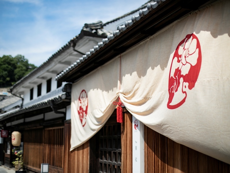 「白い暖簾に華紋の紋が映える、晴れやかな外観。青空の下、美観地区の風景に凛と佇みます。